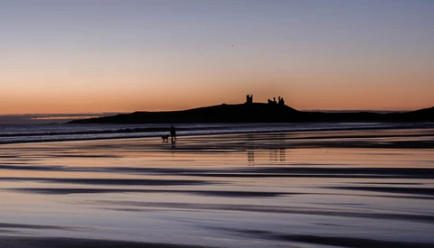Silhouetted castle ruins on a distant headland at sunset, with a lone surfer in the shallow foreground water.