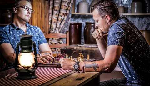 Two men playing chess by lamplight in a rustic interior setting.