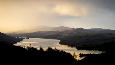 Golden-hour landscape with a still lake surrounded by forested hills and distant mountains.
