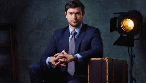 Professional man in navy suit seated next to vintage leather suitcase with studio lighting setup visible.