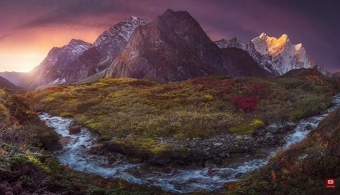 Snow-capped mountain peaks rise above a moss-covered valley with a winding blue stream at sunset.