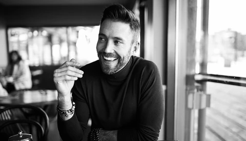 Man with beard smiling at camera in bright indoor space with windows, black and white portrait.