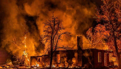 Residential structure engulfed in intense flames at night, surrounded by bare trees silhouetted against orange and black smoke.