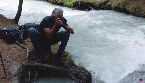 Photographer crouching by a rushing river, camera raised to eye level to capture the flowing water.