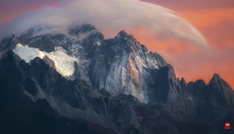 Snow-capped mountain peaks illuminated by dramatic orange and pink sunset light beneath layered clouds.