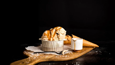 Melting ice cream scoops in a ribbed metal cup with cone and napkin on wooden board against black background.