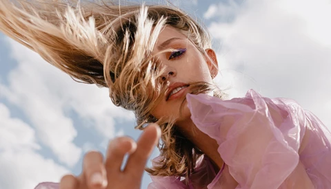 Woman in pink sheer top photographed against cloudy sky with windswept blonde hair and blue eyeshadow.