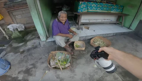 Elderly person in purple shirt preparing fresh vegetables outdoors on concrete ground.