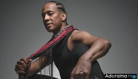 Shirtless man in profile holding a beaded pink and red shoulder scarf against gray background.
