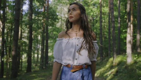 Woman in off-shoulder white blouse and denim skirt standing in a sunlit forest.