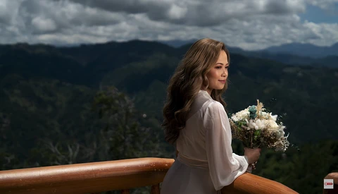 Woman in light blouse holding white flowers at wooden railing overlooking forested mountains.
