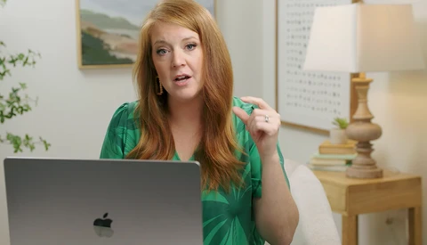 Woman with reddish-blonde hair in green shirt gesturing while speaking at laptop during video call.