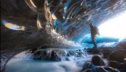 Hiker standing in an ice cave with turquoise glacial stream flowing beneath textured blue ice formations.