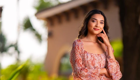 Woman in coral patterned top posing outdoors with blurred residential architecture in background.
