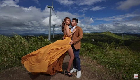 Couple posing together in a rural landscape with a wind turbine and rolling hills in the background.