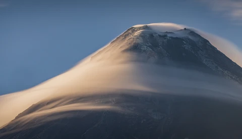 Snow-capped volcanic cone rising through layered lenticular clouds at dawn.