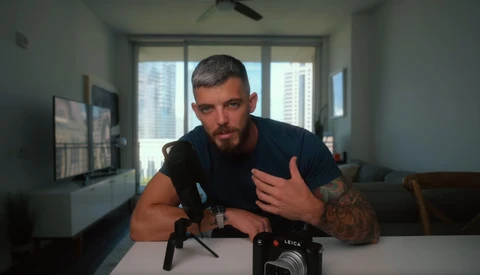 Man with tattoo gesturing while speaking at table with professional camera and microphone setup.