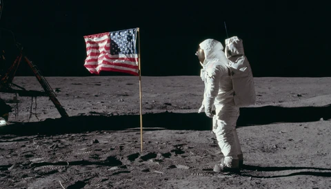 Astronaut in white spacesuit standing on lunar surface next to American flag.