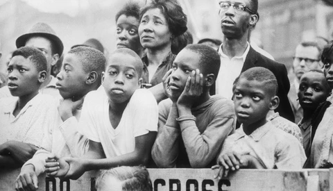Group of people of varied ages watching intently, separated by a barrier marked with partial text, in black and white.