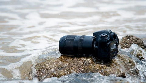 DSLR camera with telephoto lens resting on a rocky beach as waves wash over it.
