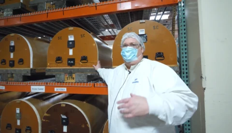 Man in white lab coat and blue mask gestures toward industrial equipment with tan-colored dome-shaped components on orange metal shelving.