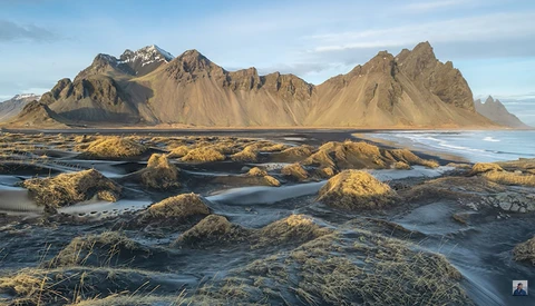 Dramatic volcanic mountain with snow-capped peaks rising behind a black sand beach strewn with moss-covered rocks and tidal pools.