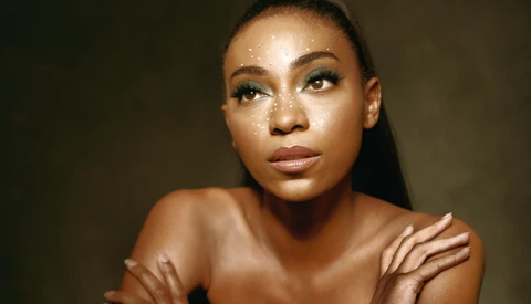 Portrait of a woman with dewy skin and defined eye makeup against a neutral brown background.