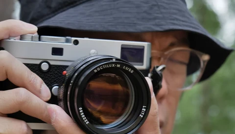 Photographer holding a Leica M rangefinder camera with a Summilux lens mounted, wearing a wide-brimmed hat.