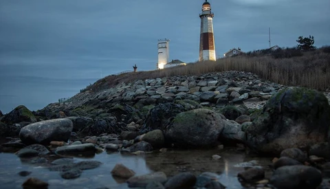 Two lighthouses on a rocky headland during twilight, with bouldery foreground and bare trees on the ridge.