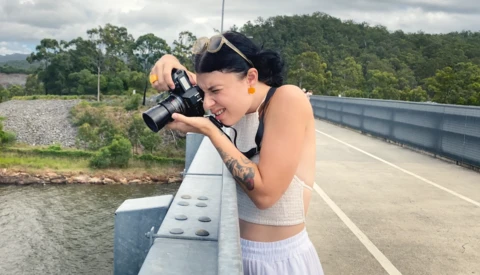 Photographer with camera on concrete bridge railing overlooking river and forested landscape.