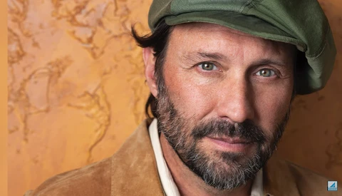 Portrait of a bearded man wearing a green cap against a textured golden-yellow background.