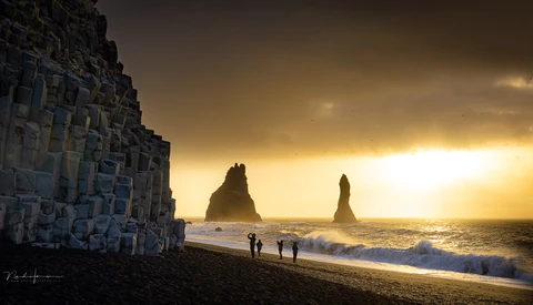 Silhouetted sea stacks rising from dark sand beach at golden hour with basalt columns on left.