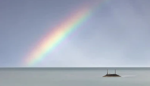 Vivid rainbow arcing over calm, misty water with a small rocky island in the distance.