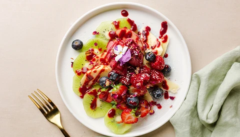 Overhead view of a plated fruit arrangement with kiwi slices, berries, and red fruit coulis on white ceramic plate.