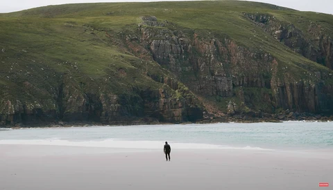 Solitary figure in dark wetsuit standing on pale sand beach beneath towering green hillside with exposed basalt columns.