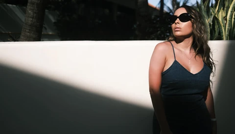 Woman in black camisole and sunglasses standing against white wall in strong sunlight.