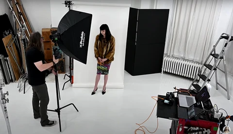 Professional studio setup with photographer operating camera on tripod, subject in brown jacket and patterned skirt positioned under softbox lighting, black reflectors and additional equipment visible.