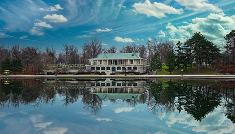 White plantation-style house reflected in still water with bare trees and evergreens along the shoreline.
