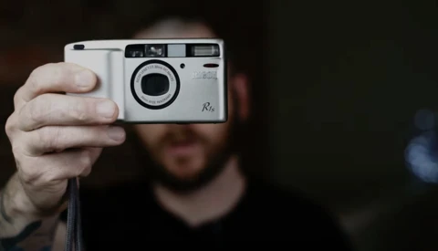 Photographer holding a white point-and-shoot film camera with a large circular lens element, displaying the camera toward the viewer.