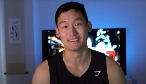 Young man in black sleeveless shirt smiling at camera in blue-lit room with monitors in background.
