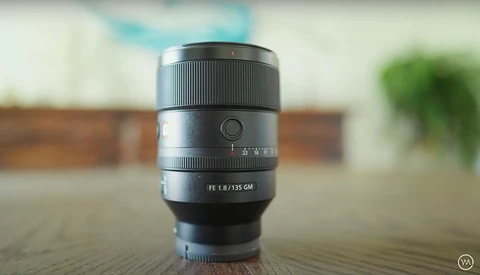 Sony FE 1.8/135 GM lens displayed upright on a wooden surface with blurred interior background.