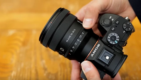 Hand holding a mirrorless camera with attached wide-angle zoom lens against warm wooden background.