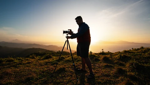 Silhouetted videographer operating camera on tripod during golden hour with mountains in background.