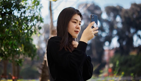 Woman holding smartphone horizontally to capture a photograph, with blurred urban landscape and green foliage in background.