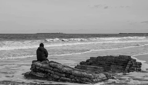 Person sitting on a textured rock formation in shallow ocean waves under an overcast sky.