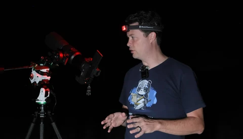Photographer wearing headlamp demonstrates camera setup with red-lit telescope equipment in dark studio.