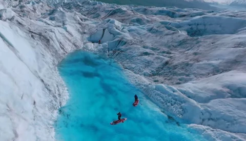 Two kayakers paddling through a turquoise glacial meltwater lake surrounded by towering white ice walls and blue crevassed glacier.