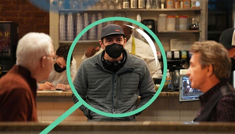 Man wearing a cowboy hat and black face mask stands behind a counter, highlighted with a green circle overlay.