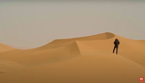 Solitary figure in dark clothing standing on a massive sand dune under hazy sky.