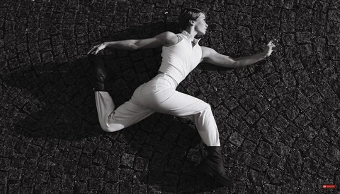 Male dancer in white outfit performing a dynamic leap against textured dark wall.